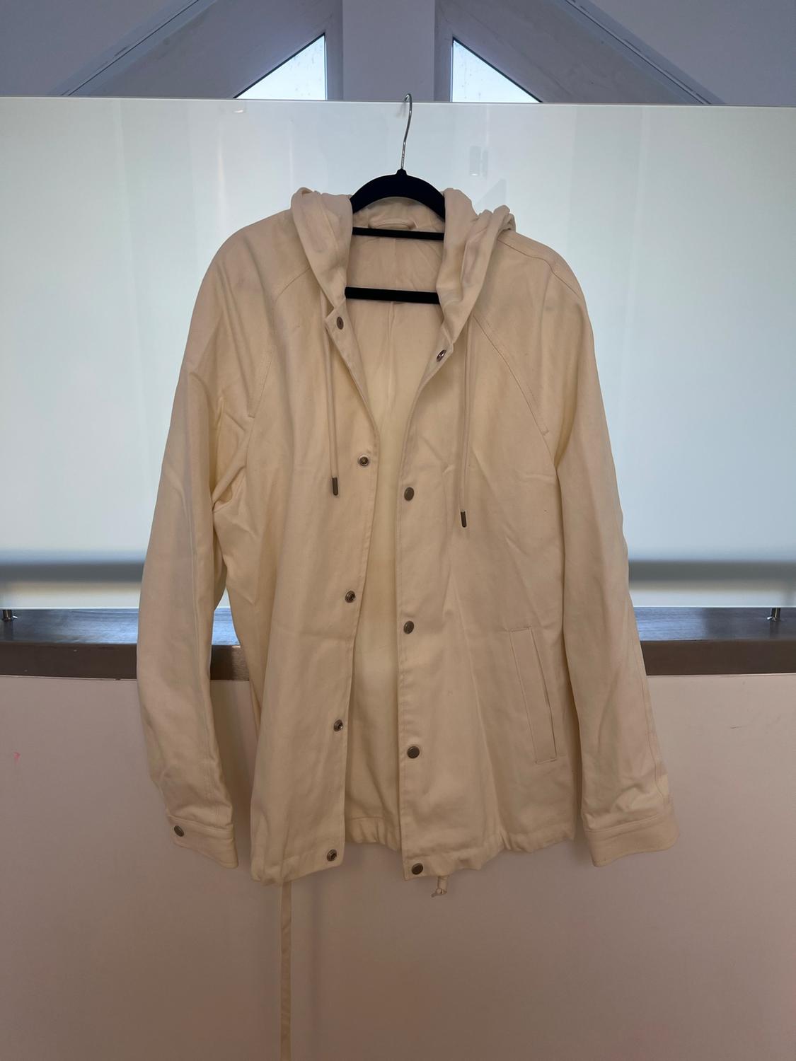 Cream-coloured hooded jacket with front buttons, hanging on a black hanger in front of a translucent glass window.