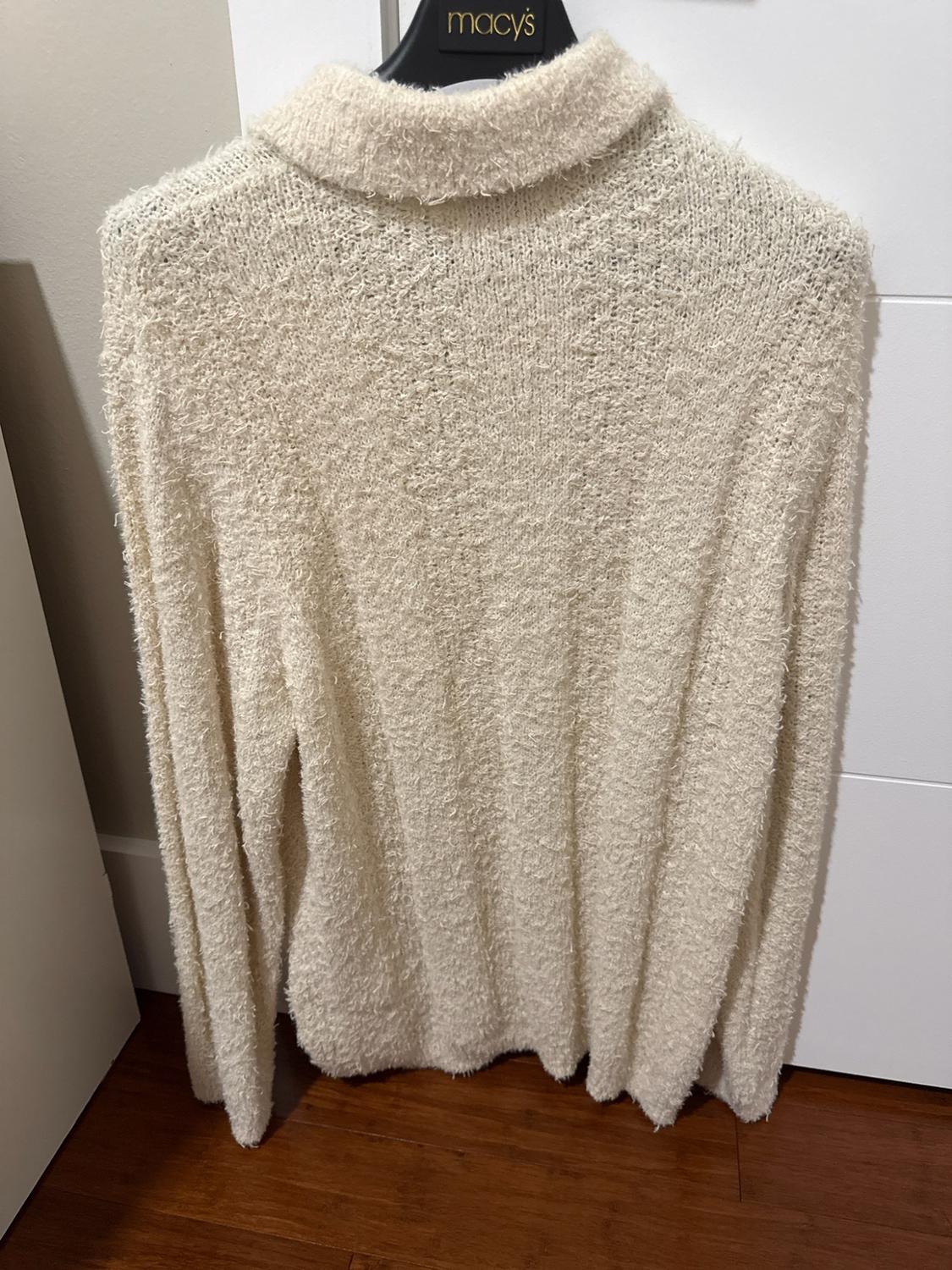 Cream textured-knit sweater with a turtleneck on a black hanger featuring the text “macy’s,” hanging on a white door above a wooden floor.