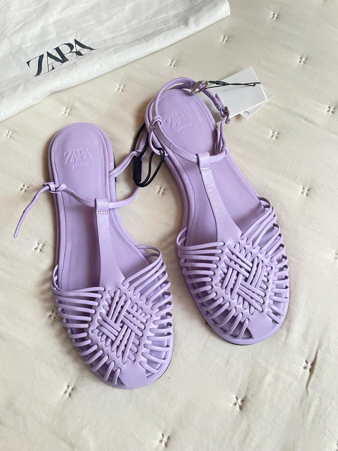 Lilac sandals with a braided design on a beige surface. In the background, a white bag with the "ZARA" logo.