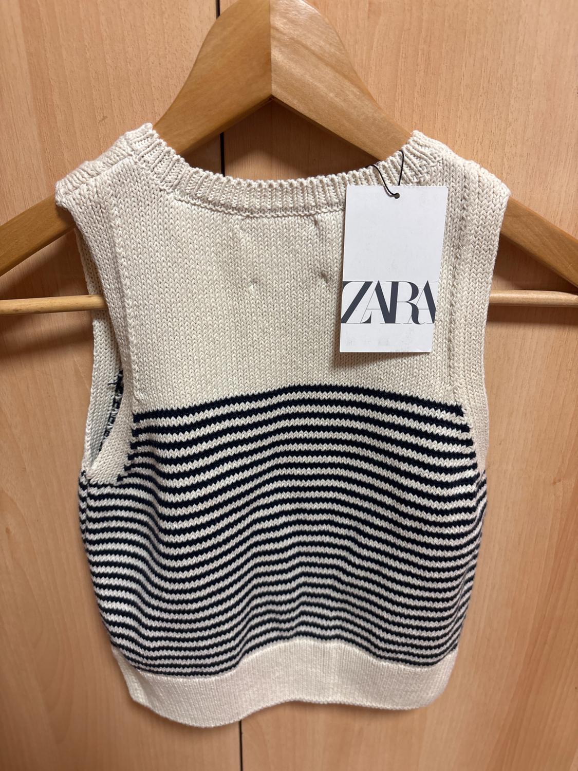 Cream knitted waistcoat with dark horizontal stripes, on a wooden hanger against a wooden background. Label with the text ZARA.