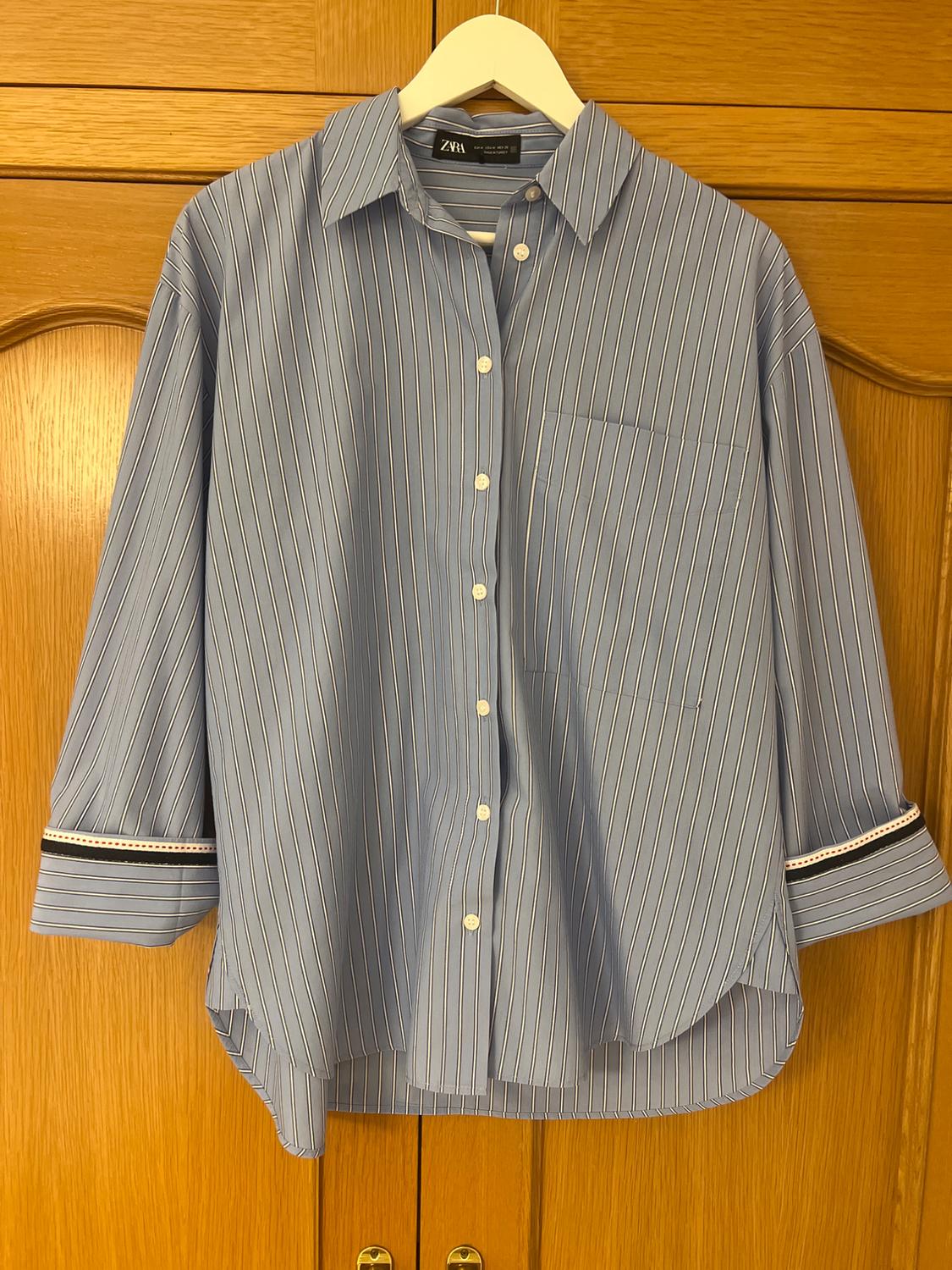 Blue shirt with white stripes, featuring a front button fastening and visible ZARA label. Hanging on a white hanger against a wooden door background.