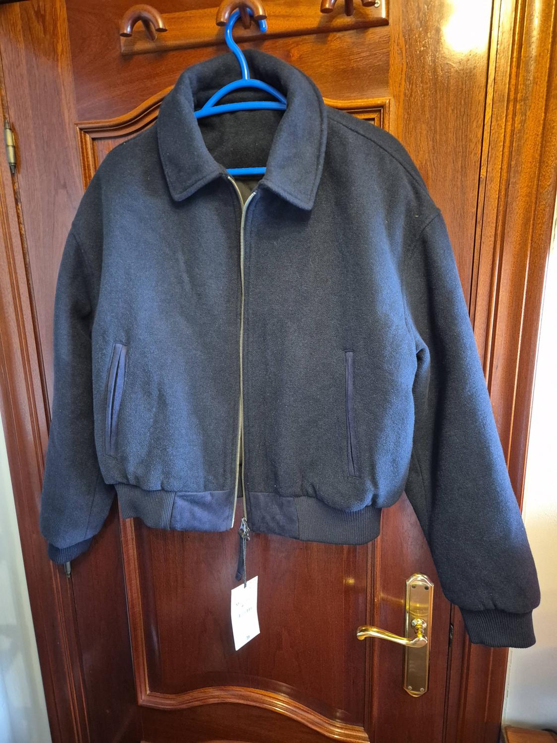Navy blue jacket with a zip and a hanging tag, on a blue hanger over a wooden door with a gold handle.