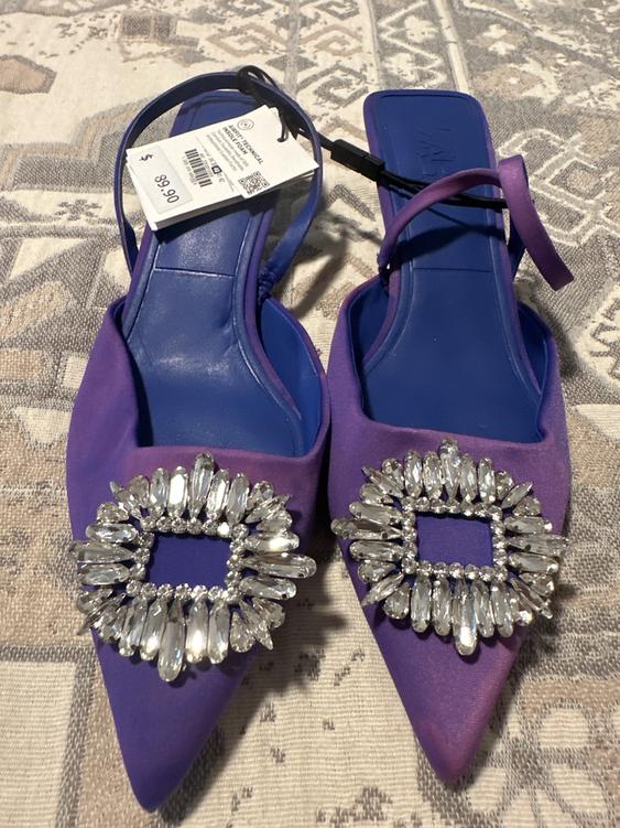 Purple high heels with rhinestone embellishments on the toe, price tag of $89.90, on a geometric patterned rug.