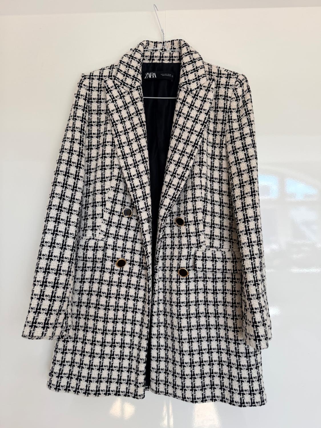Black and white checked coat hanging on a hanger against a white background. It has dark buttons and a label bearing the text ZARA.
