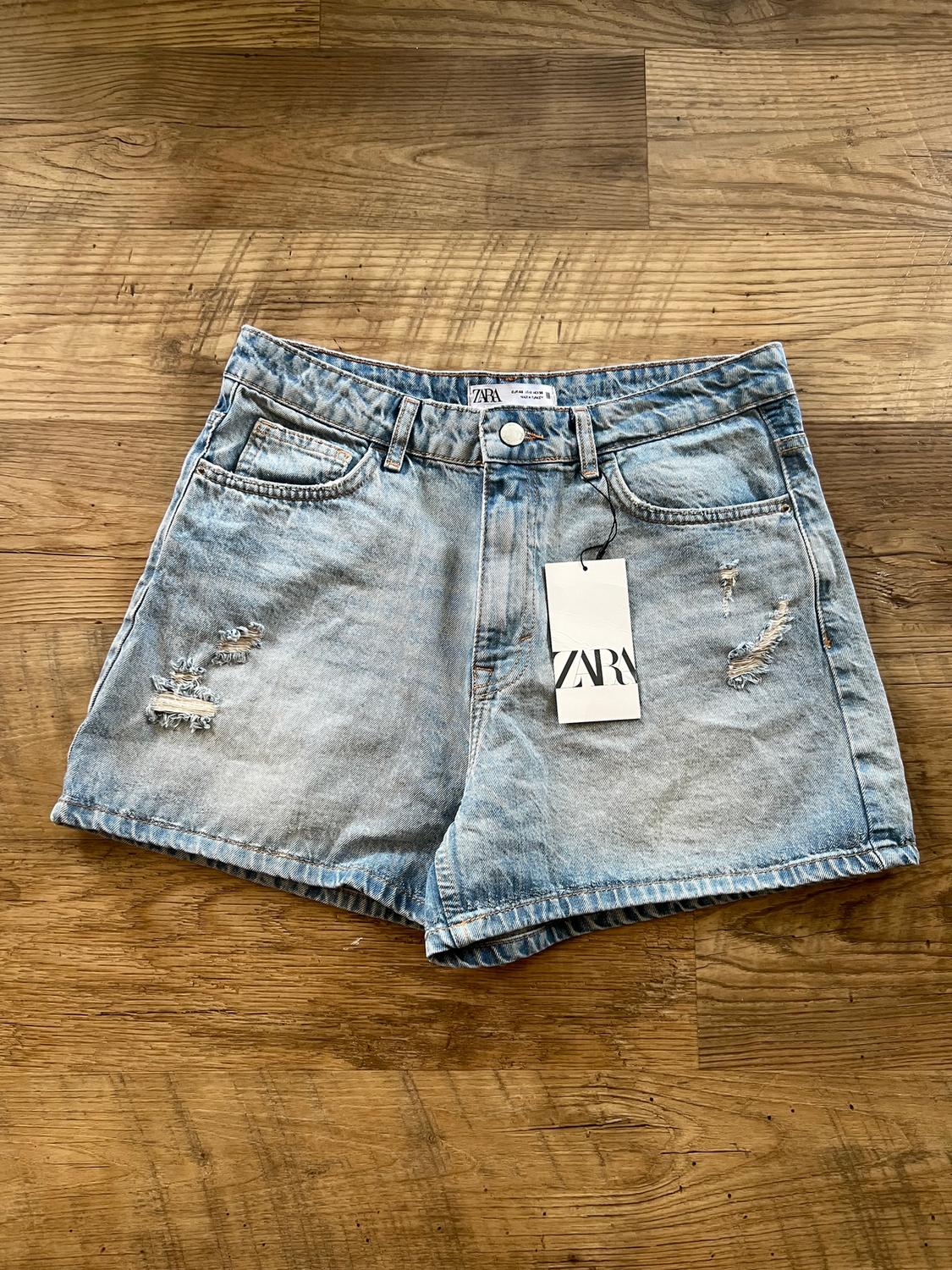 Light blue denim shorts with distressed details and a Zara tag on a wooden floor.