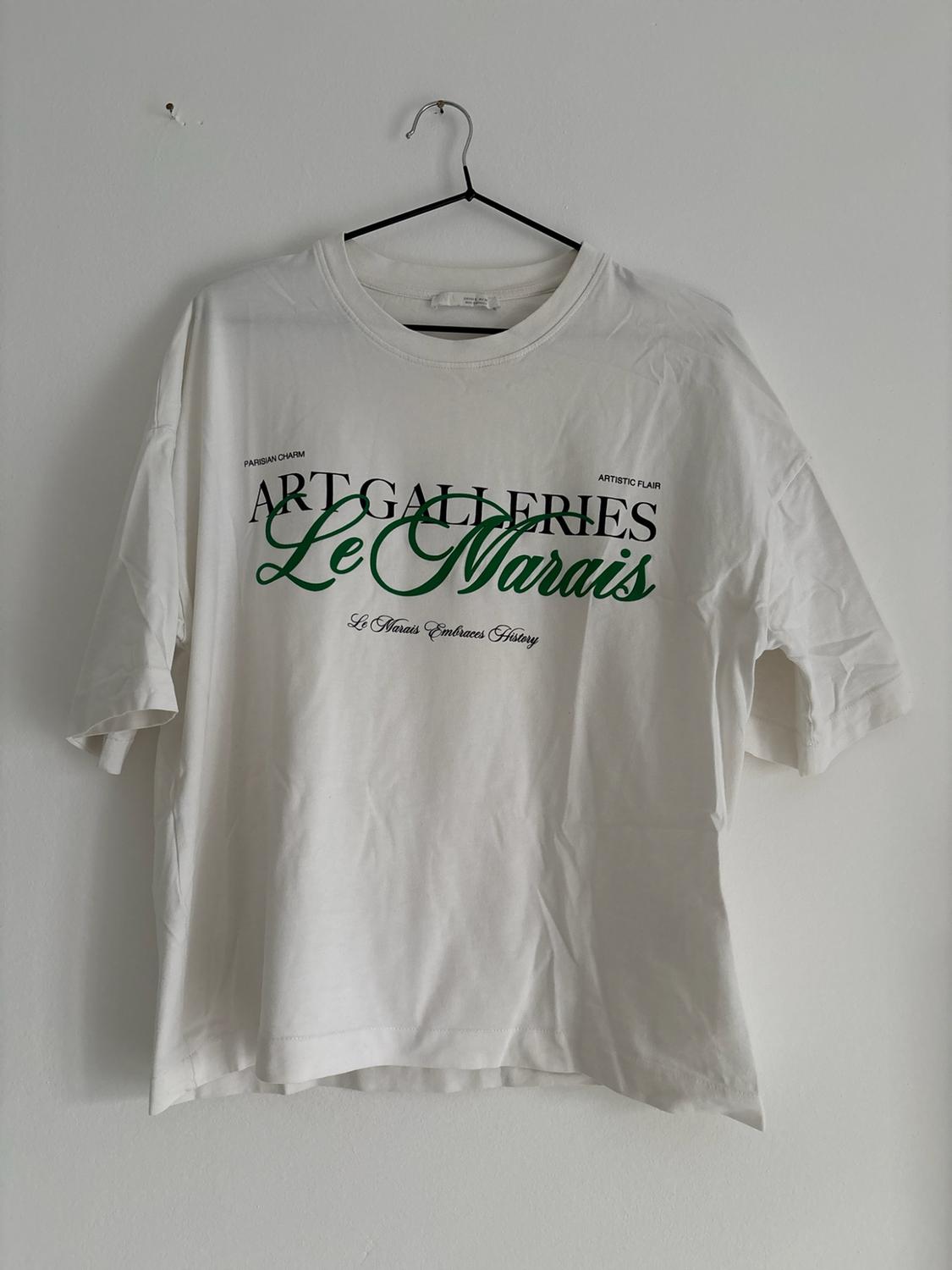 Hanging white t-shirt with text: PARISIAN CHARM, ARTISTIC FLAIR, ART GALLERIES, Le Marais, Le Marais Embraces History.