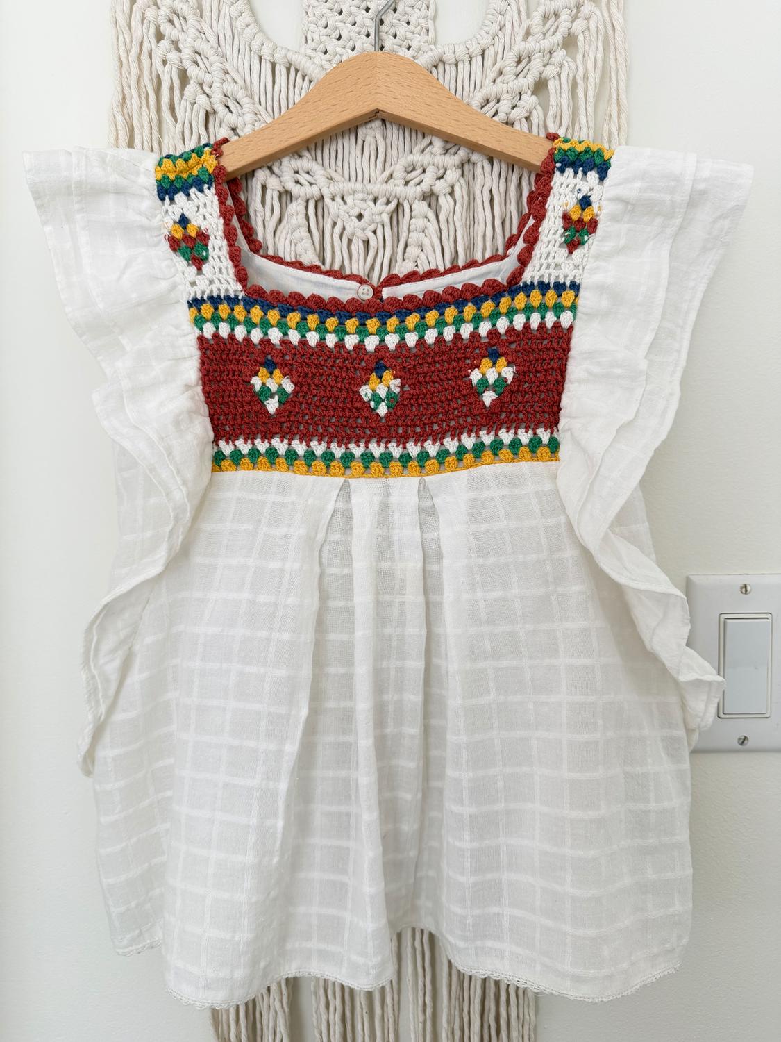 White blouse with colorful embroidery on the chest, hanging on a wooden hanger against a beige macrame background.