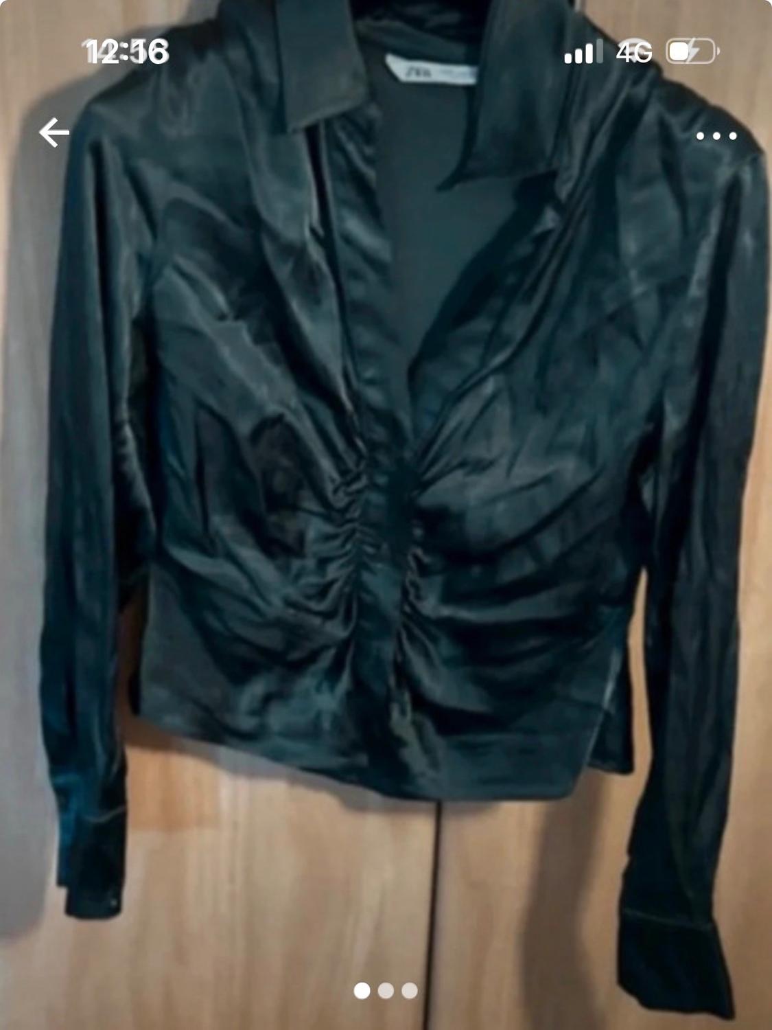 Black satin blouse with ruched detailing in the centre, hanging on a wooden door. Phone screen visible with time 12:36.