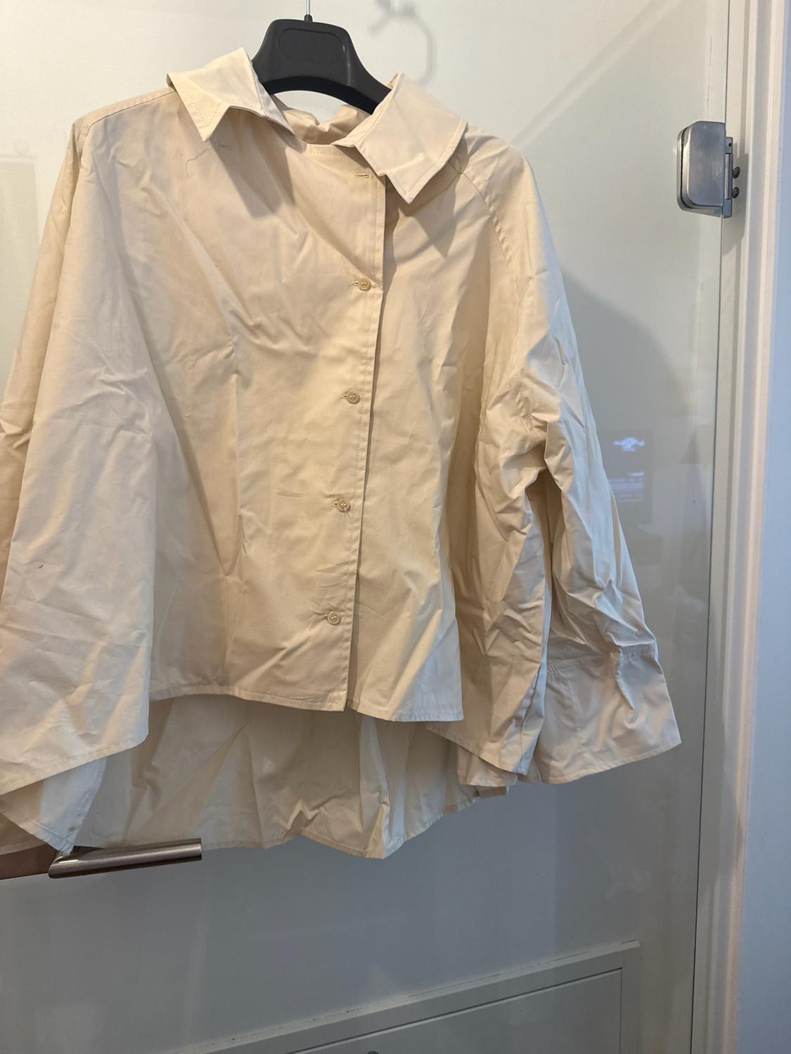 Beige long-sleeved shirt with buttons and an asymmetric hem, hanging on a black hanger over a glass door.