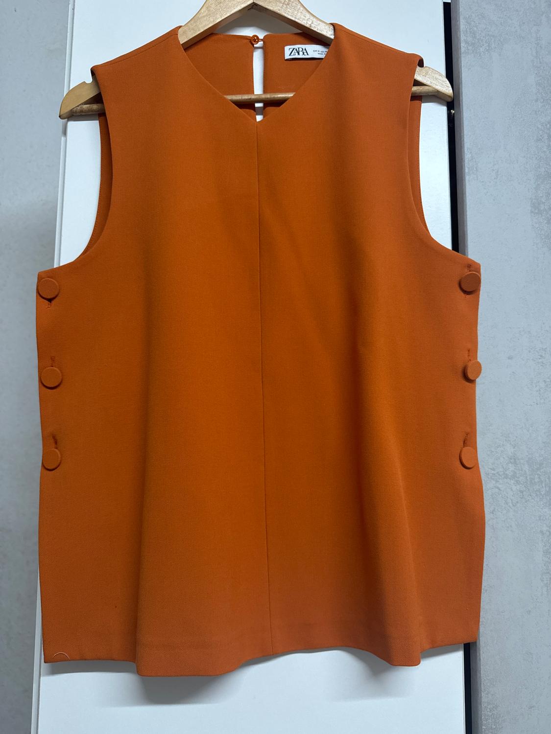 Orange sleeveless blouse hanging on a wooden hanger, with decorative buttons on the sides. Visible tag: Zara.
