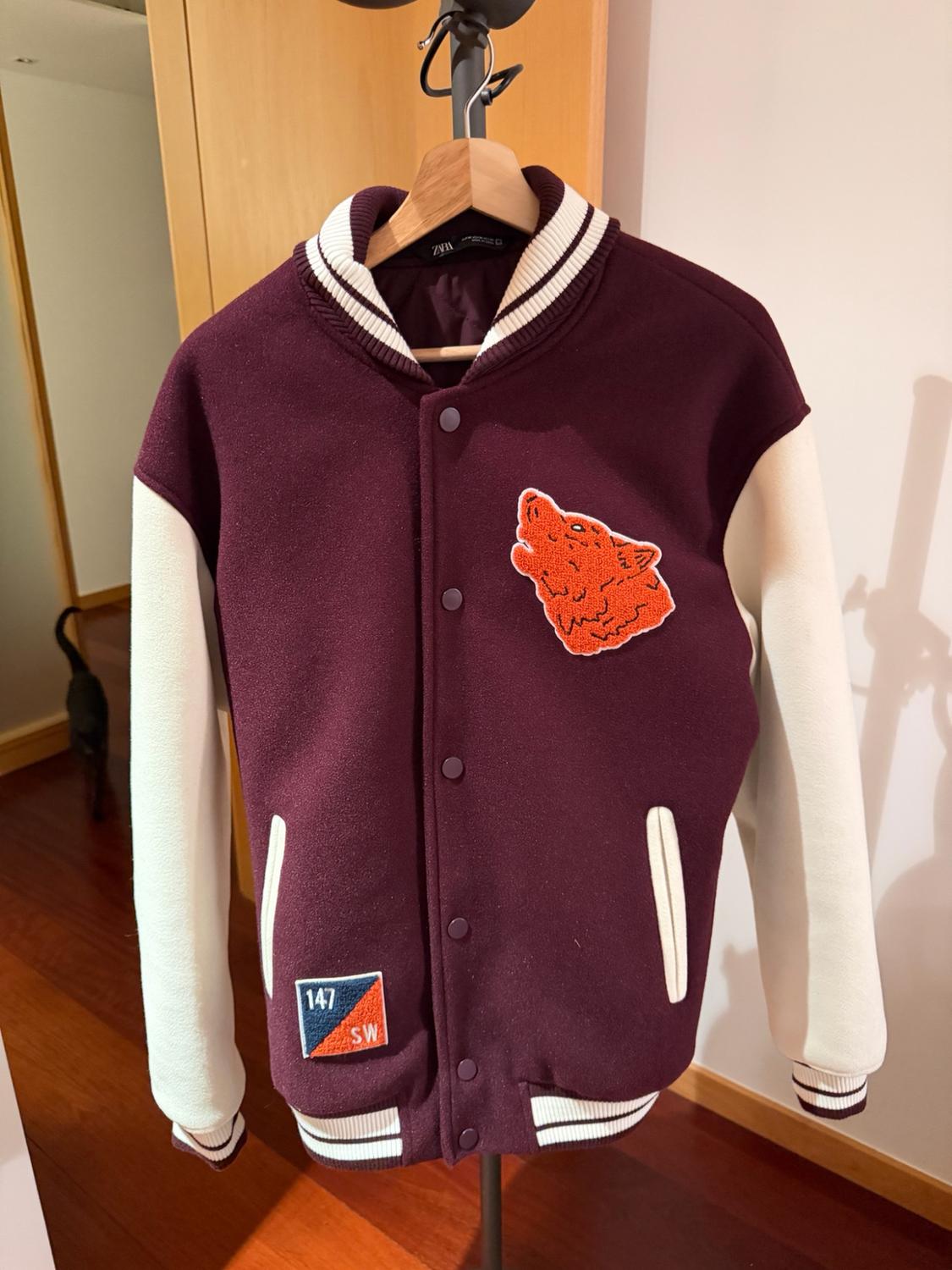 Γρενά και λευκό varsity jacket σε κρεμάστρα. Πορτοκαλί μπάλωμα με λύκο και το κείμενο 147 SW. Ετικέτα ZARA. Εσωτερικό με γάτα στο βάθος.