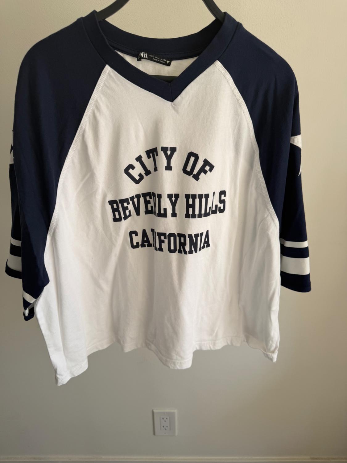 White and blue T-shirt with raglan sleeves and "City of Beverly Hills California" text on the front, hanging on a hanger.