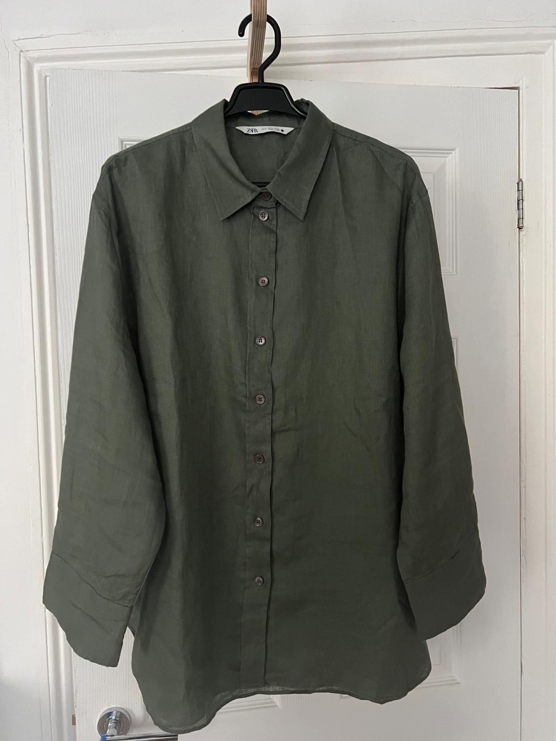 Olive green long-sleeved button-up shirt, hanging on a black hanger against a white door. The label reads ZARA.