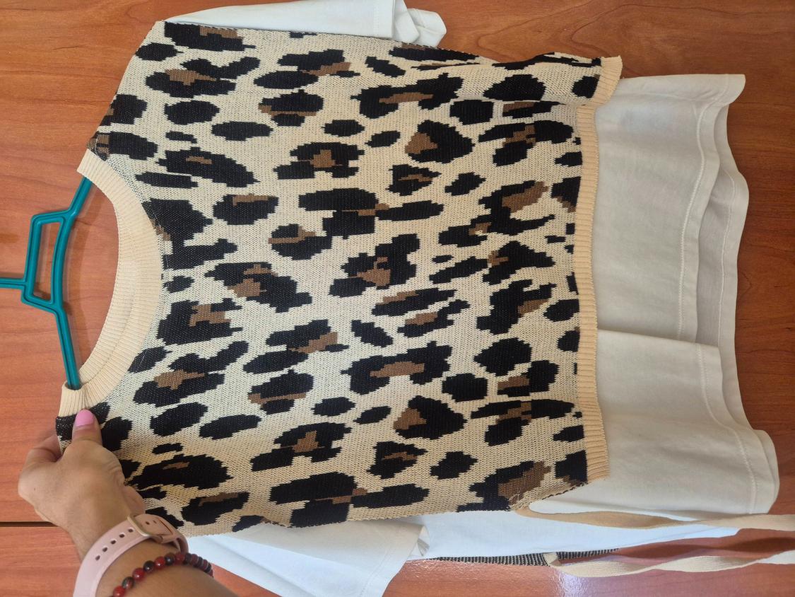 Hand holding a leopard-print knit top on a hanger over a white t-shirt, resting on a wooden table.