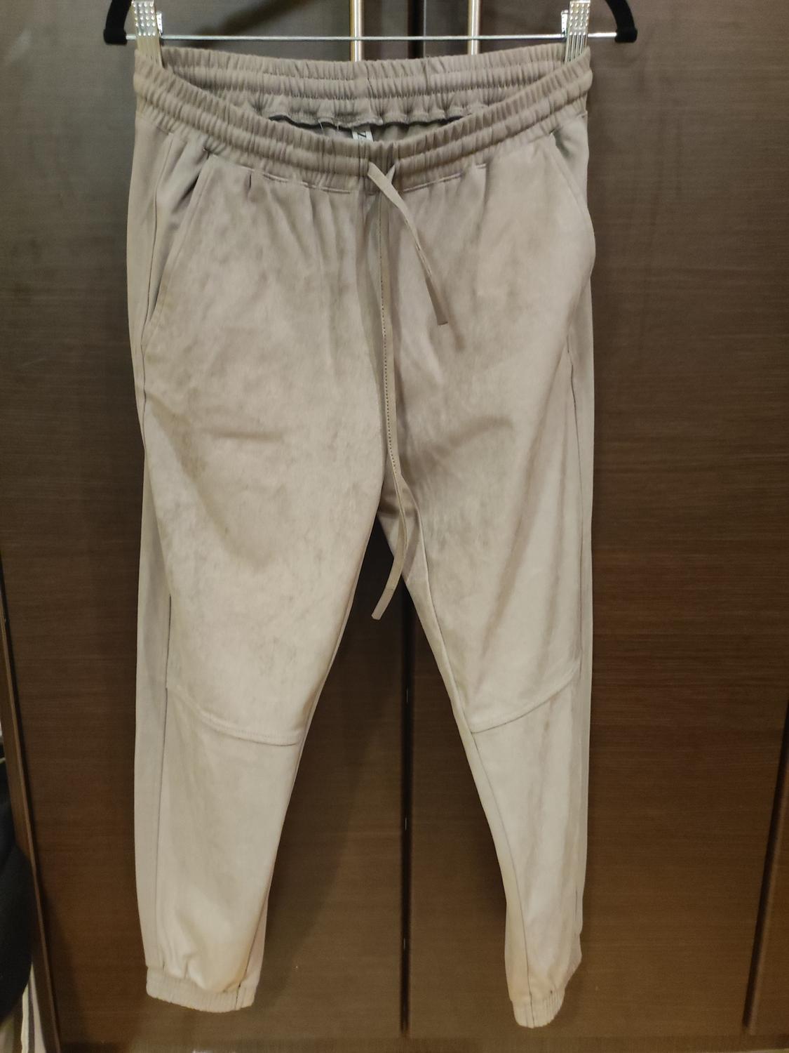 Beige jogger trousers hanging on a hanger, with an elasticated waist and adjustable drawstring, dark wooden background.