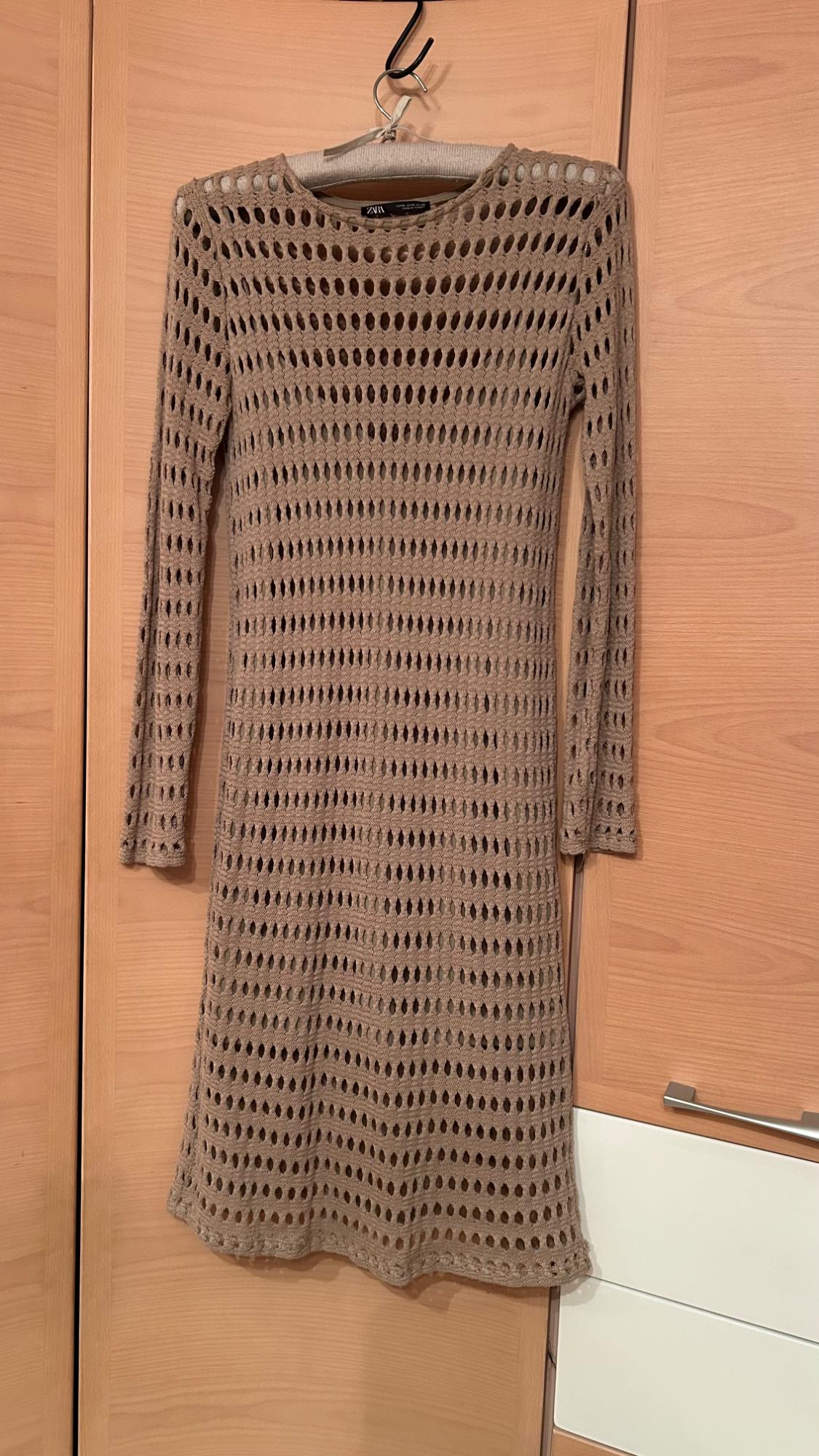 Beige open-knit long-sleeved dress hanging on a hanger in front of a light wood wardrobe. Label with the text ZARA.