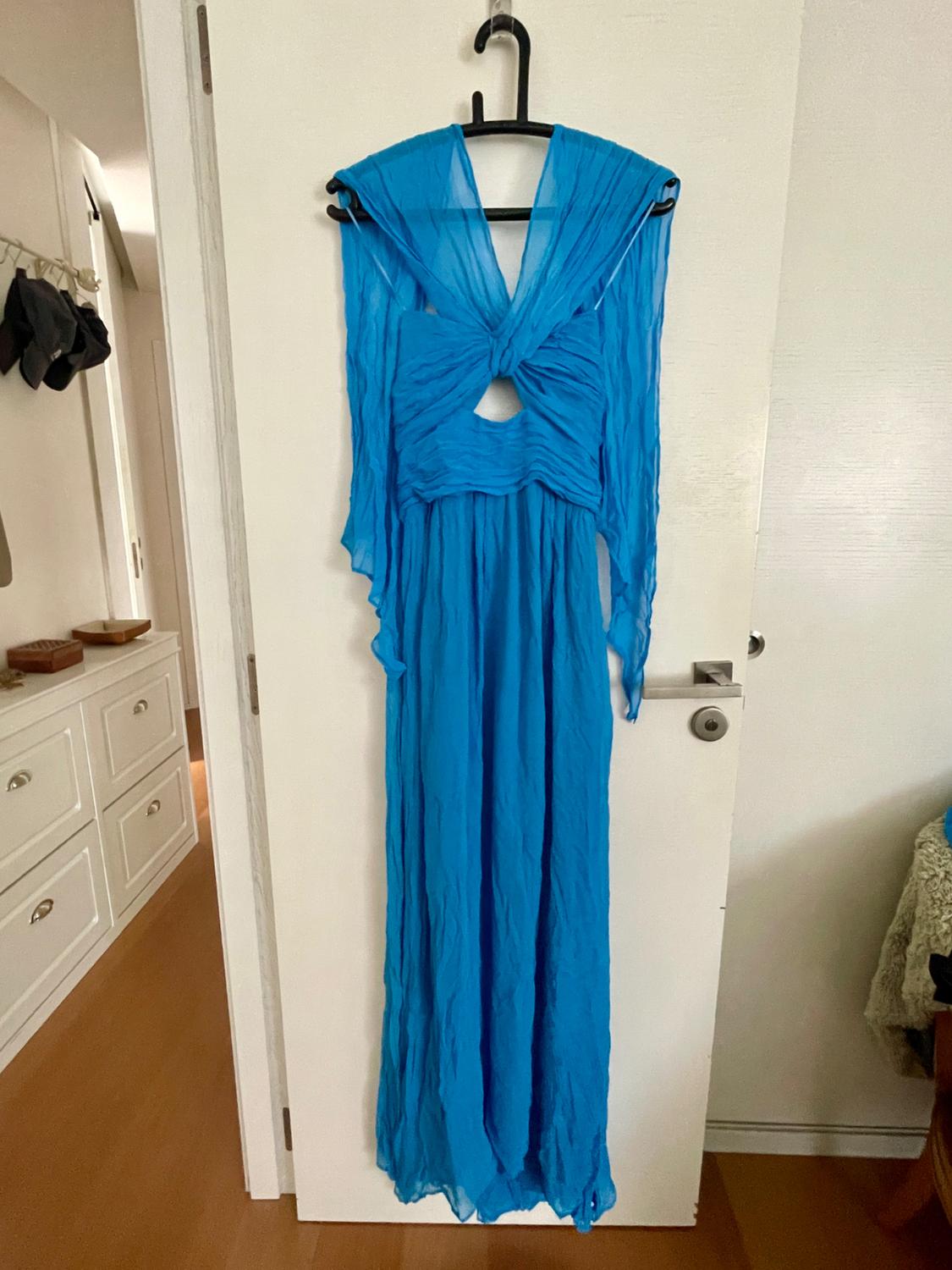 Flowing blue maxi dress with a V-neck, front knot and sheer sleeves, hanging on a white door in a relaxed home setting.