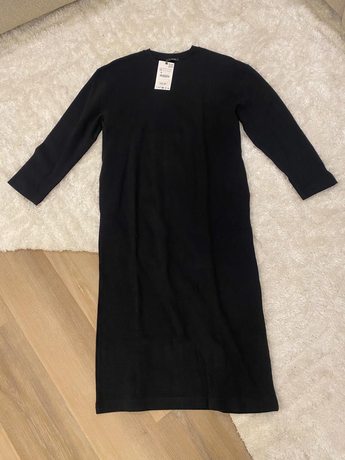 Long-sleeved black dress on a white rug and wooden floor. Visible tag on the collar with size and price details.
