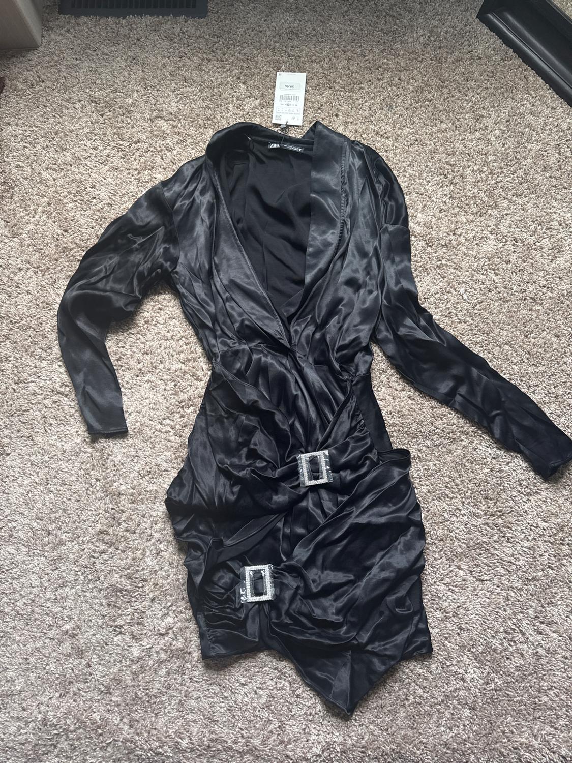 Short black satin dress, long sleeves, V-neck, and draped skirt with two sparkly buckles on a beige rug. ZARA label.