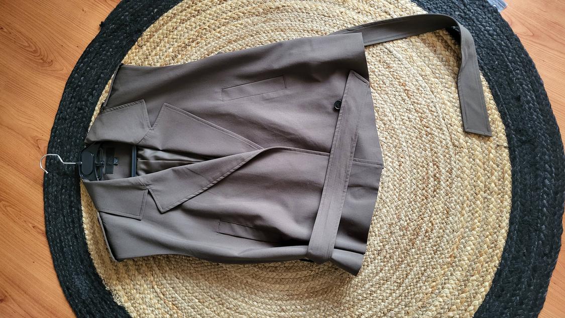 Brown belted jacket on a round jute and black rug, hung on a hanger over a light wooden floor.