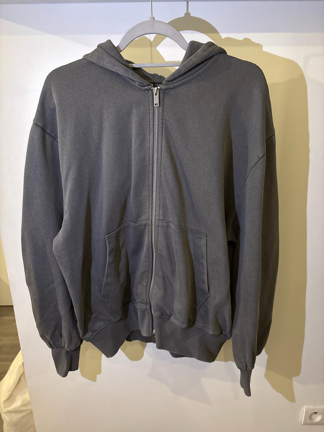 Dark grey hooded sweatshirt with zip and pockets, hanging on a hanger against a light wall with cast shadows.