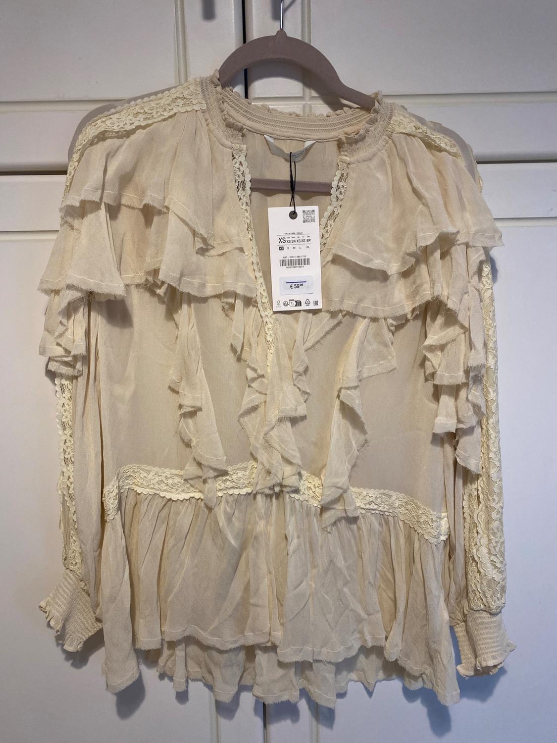 Beige long-sleeved blouse with ruffles and lace, hanging on a hanger in front of a white wardrobe. Label with text XS and €59.95.