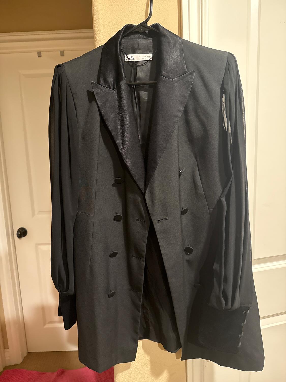 Black double-breasted jacket hanging on a hanger, with satin lapels and long sleeves, in an interior with white doors.