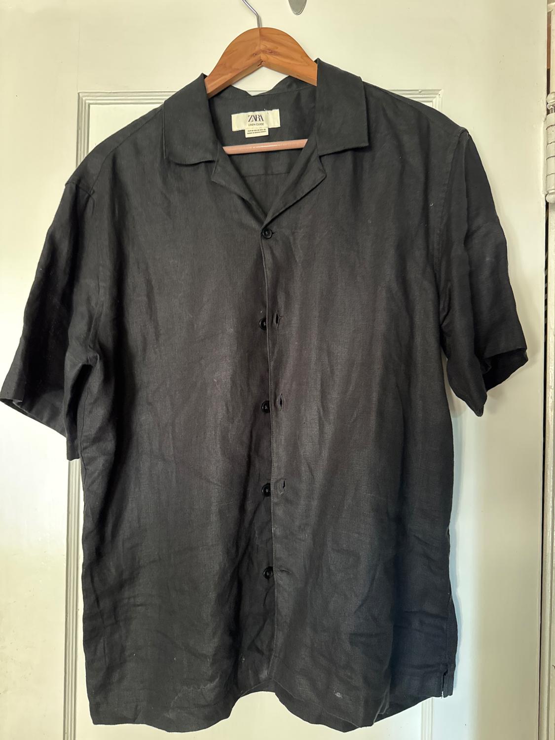 Black short-sleeved button-up shirt, hanging on a wooden hanger on a white door. The inner label reads ZARA.