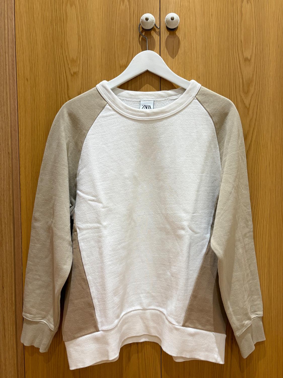 Long-sleeved sweatshirt in beige and white tones, hanging on a white hanger over a light wooden door. Visible label: ZARA.