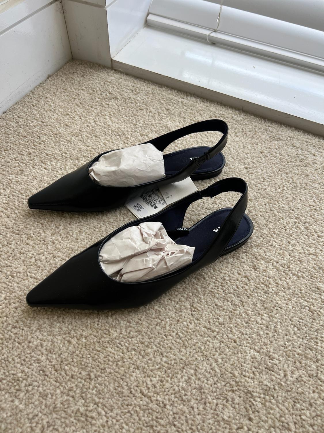 Black pointed slingback flats with paper inside, on a beige carpet next to a white skirting board. Visible text: ZARA.