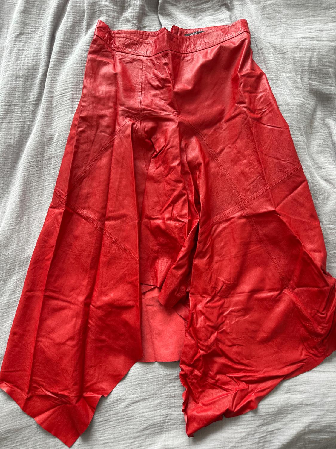 Red pleated leather skirt, laid flat on a grey fabric surface.
