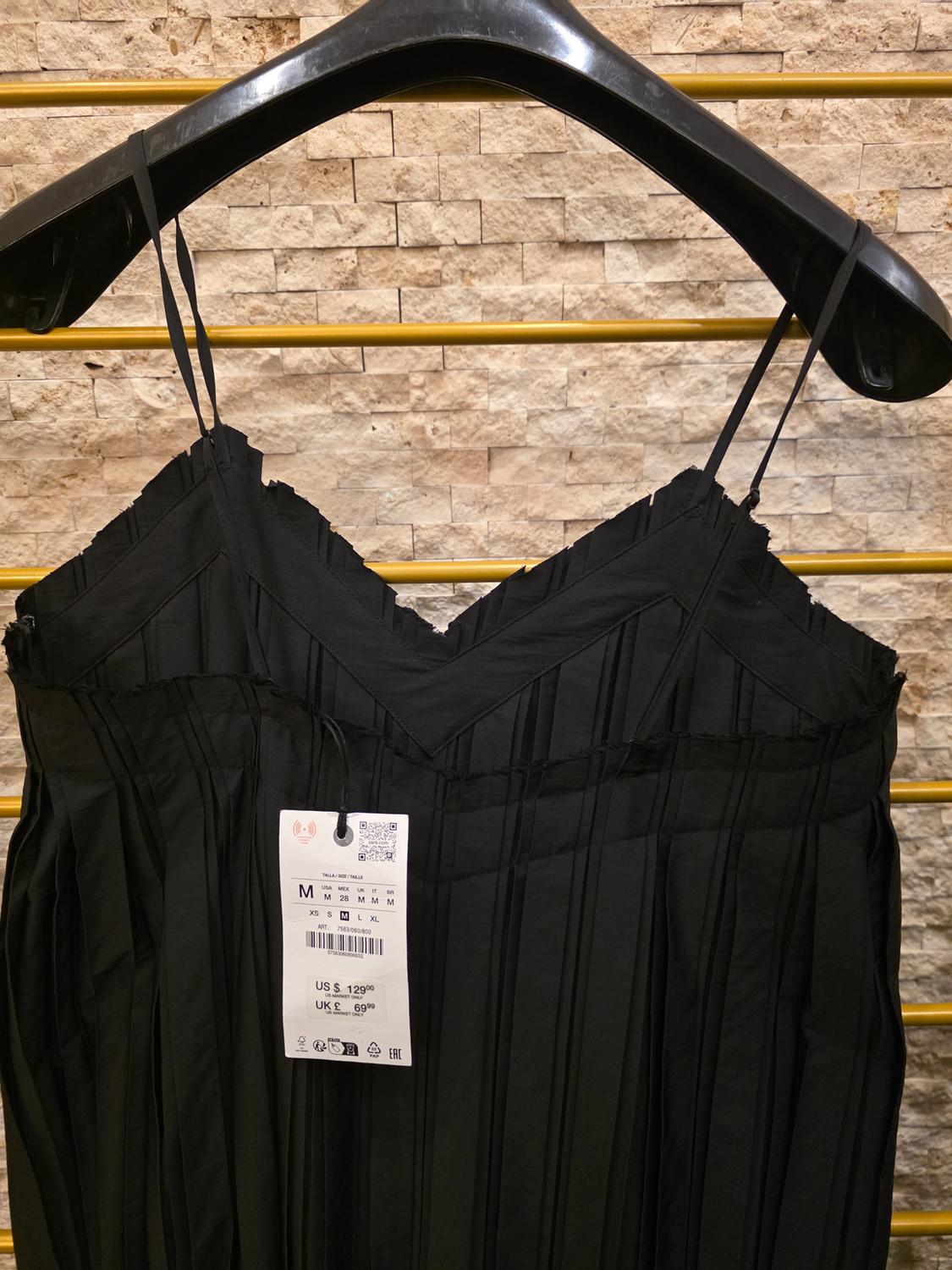Black spaghetti strap dress on a black hanger, with a visible price tag. Light brick wall background with golden bars.