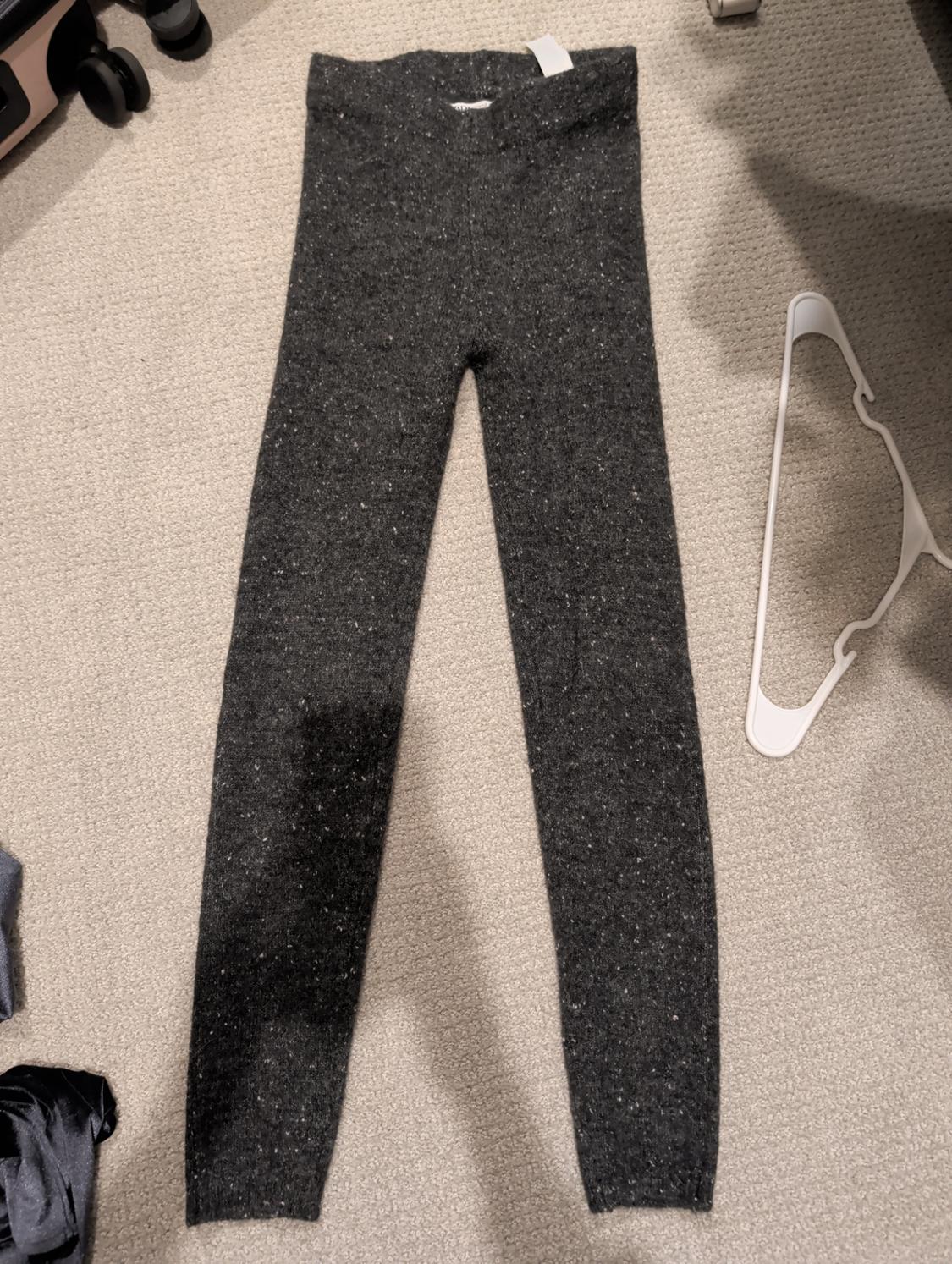 Grey flecked wool trousers on a beige rug, next to a white clothes hanger and a black suitcase.