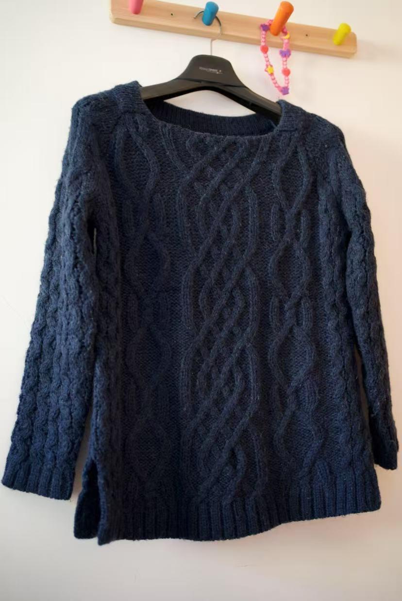 Navy blue chunky knit jumper with a cable knit design, hanging on a black hanger on a colourful wall hook.