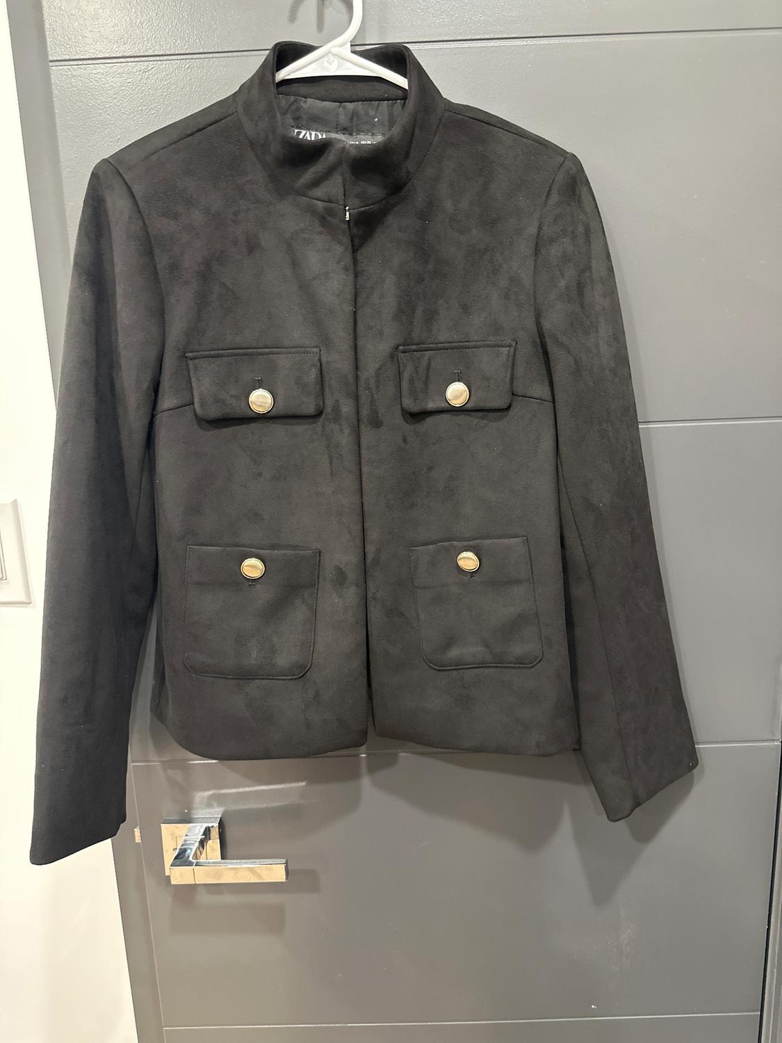 Black suede jacket with a high collar and four pockets with gold buttons, hanging on a hanger over a gray door.
