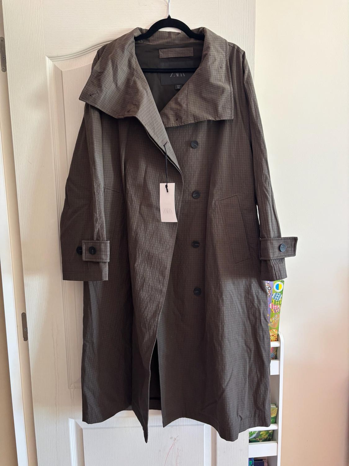 Long, dark, wide-collared trench coat with a check pattern, hanging on a white door. Label with the text ZARA.