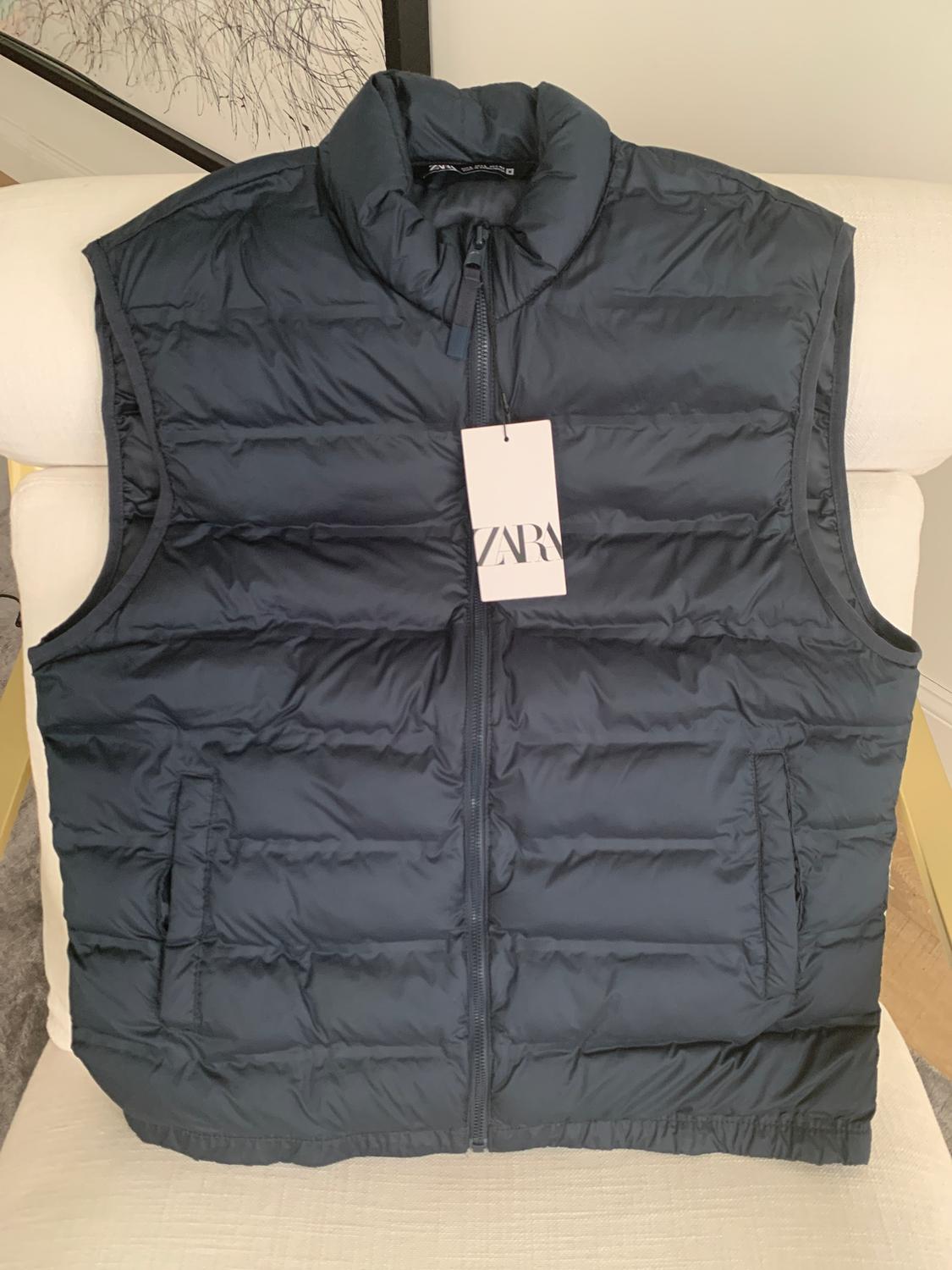Black padded gilet with a zip and high collar over a white chair. Hanging tag with the text "ZARA".