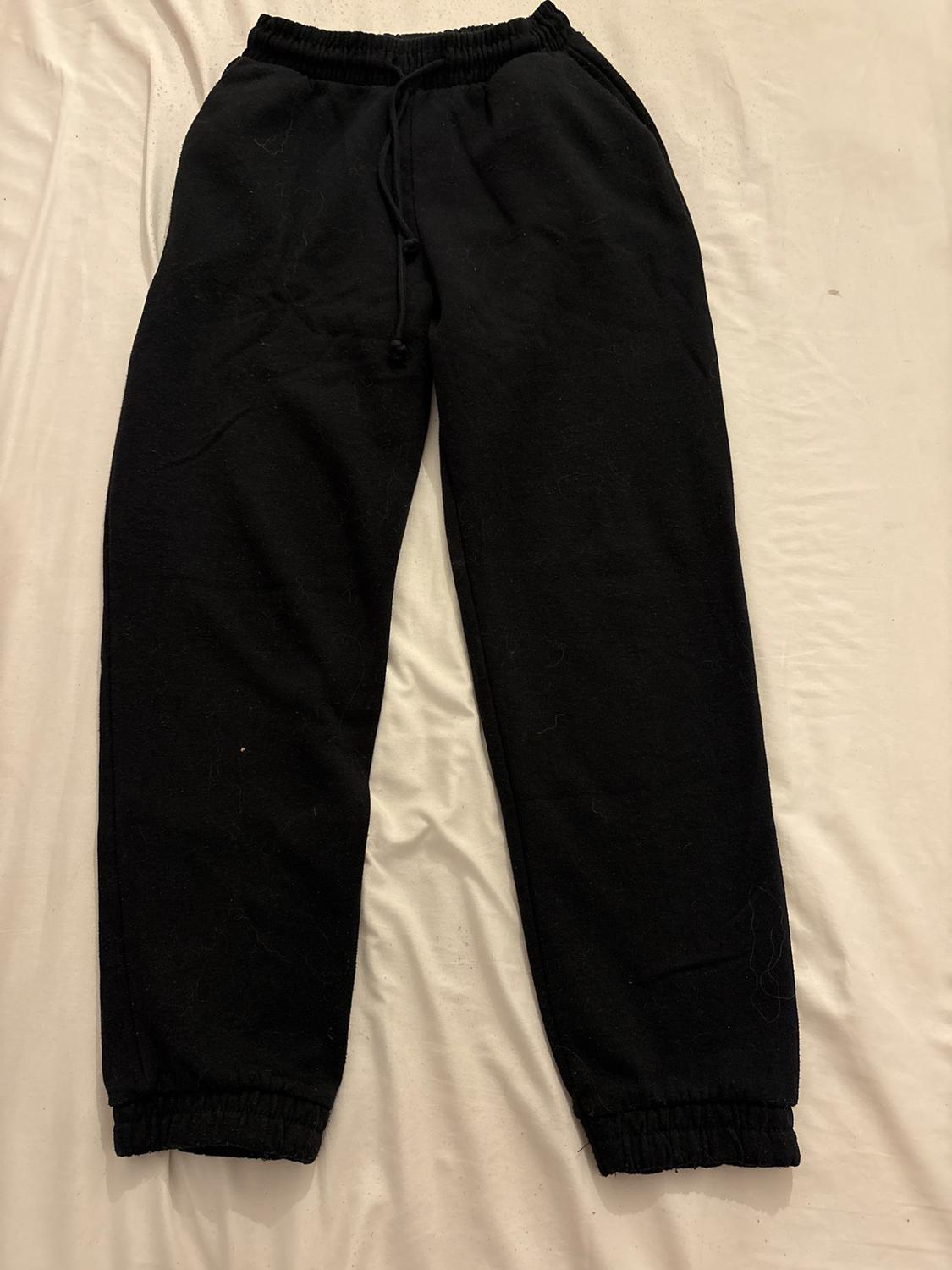Black jogging bottoms with an elasticated waist and cuffs and an adjustable drawstring, on a rumpled white sheet. Casual style.