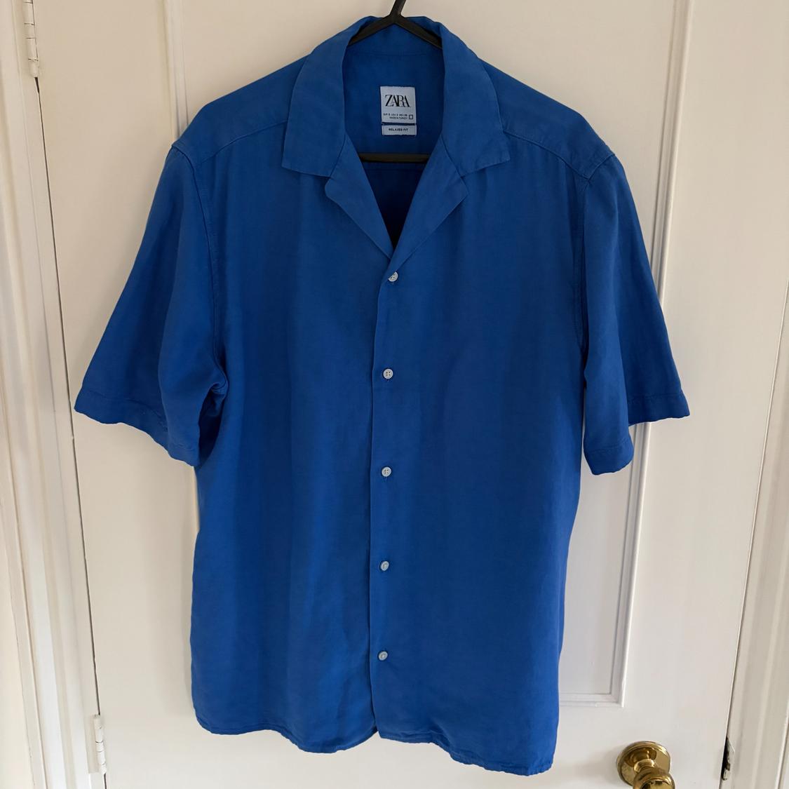 Blue short-sleeved shirt hanging on a black hanger over a white door. Label visible with the text "ZARA".