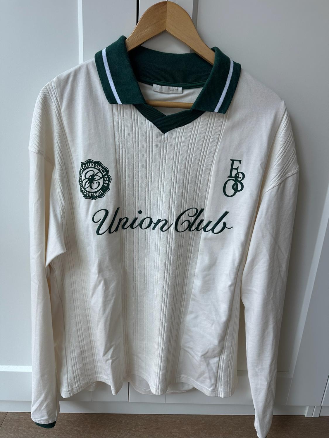 Ribbed white polo with a green collar. Texts: CLUB SINCE 1989, TIMELESS MOVEMENT, Union Club. Hanging on a hanger.