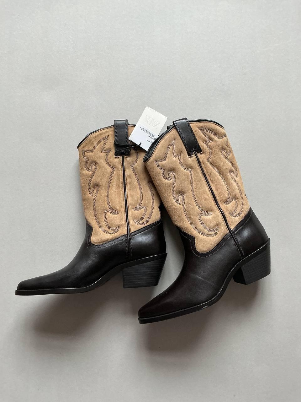 Black leather and beige suede cowboy boots with embroidered design, low heel, and hang tag on a light grey background.