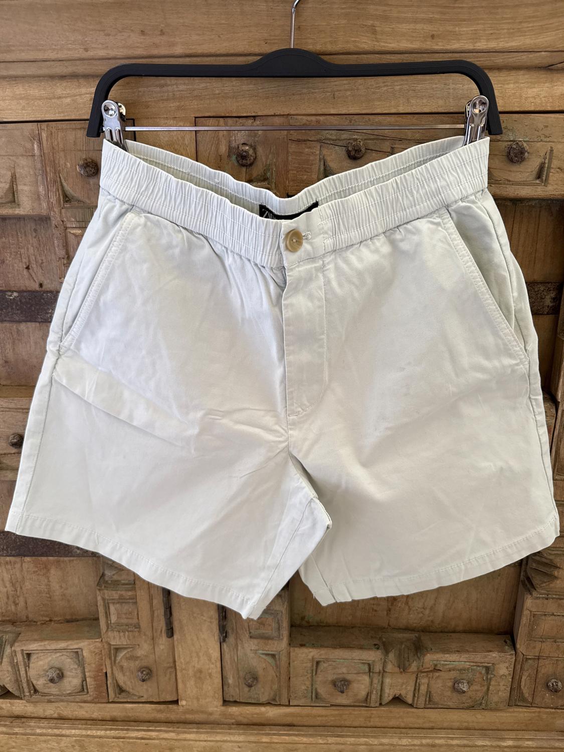 Beige shorts hanging on a black hanger against a carved wooden background. Elasticated waist and visible front button.