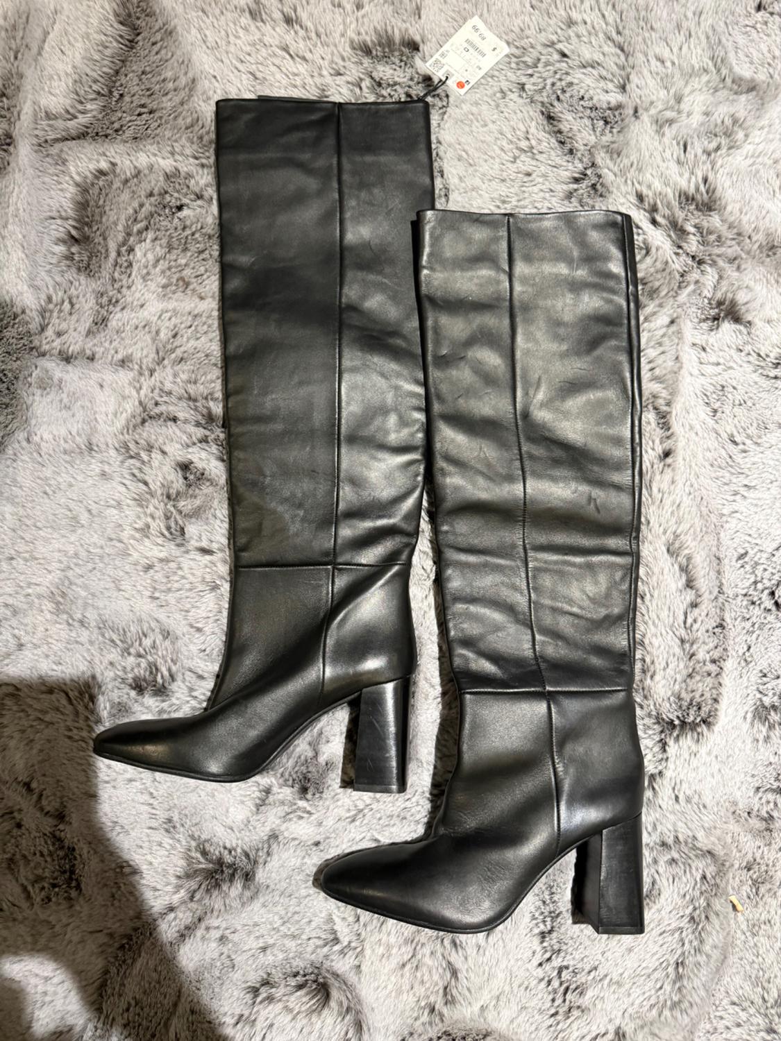 Black chunky-heeled tall boots on a gray shag rug. Visible tag on the top of one boot.