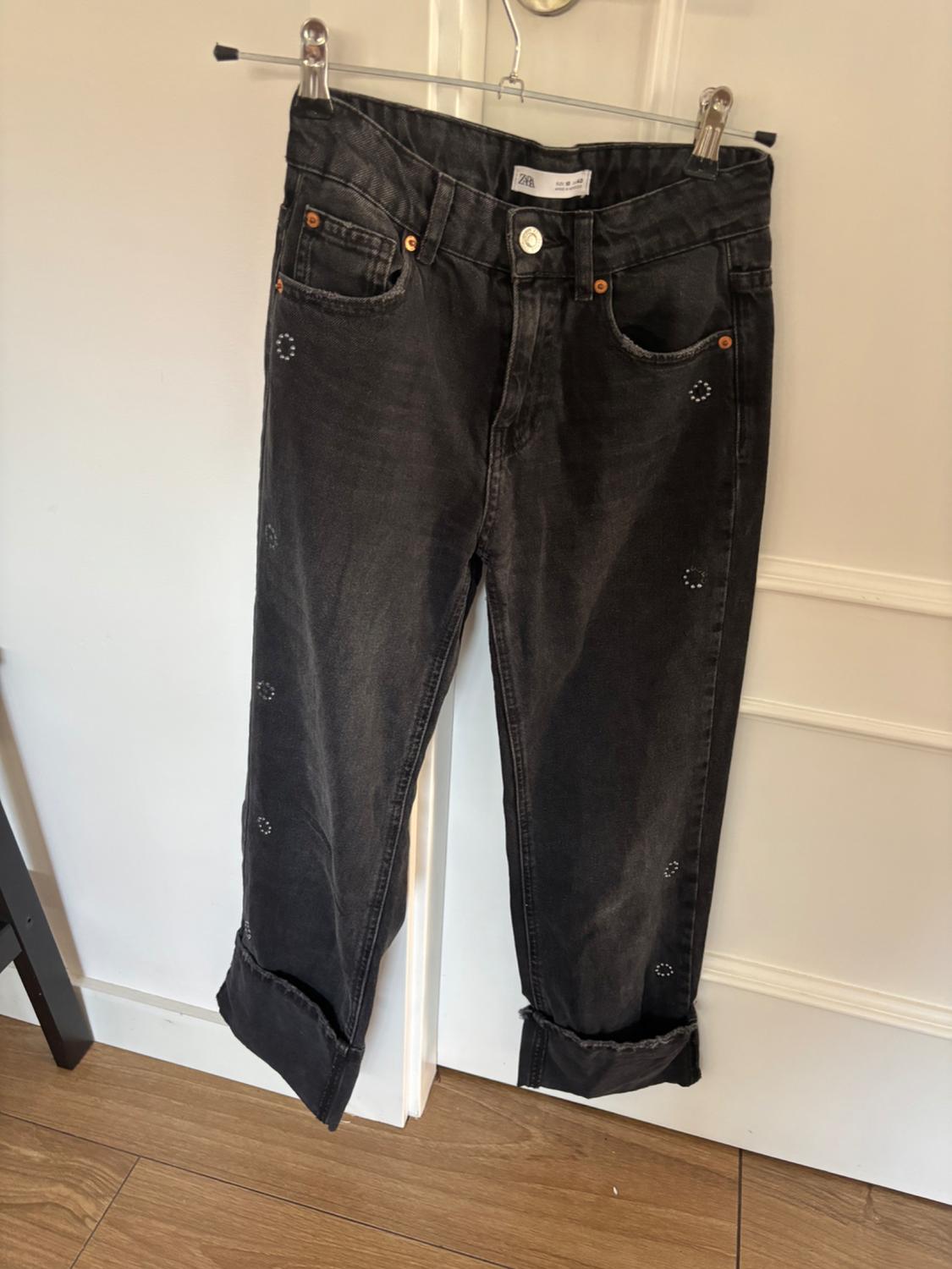 Black jeans hanging on a clothes hanger, with stud detailing on the pockets and legs. White door background and wooden floor.