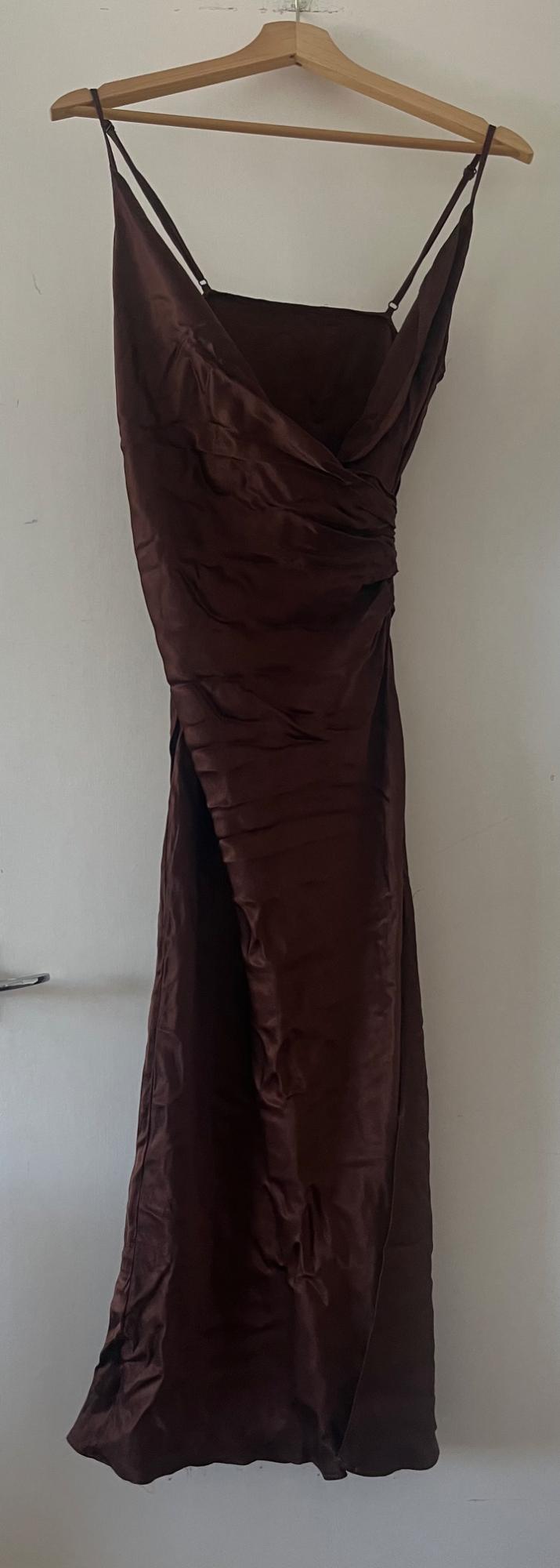 Brown satin maxi dress with thin straps and draped detail, hanging on a wooden hanger against a white door.