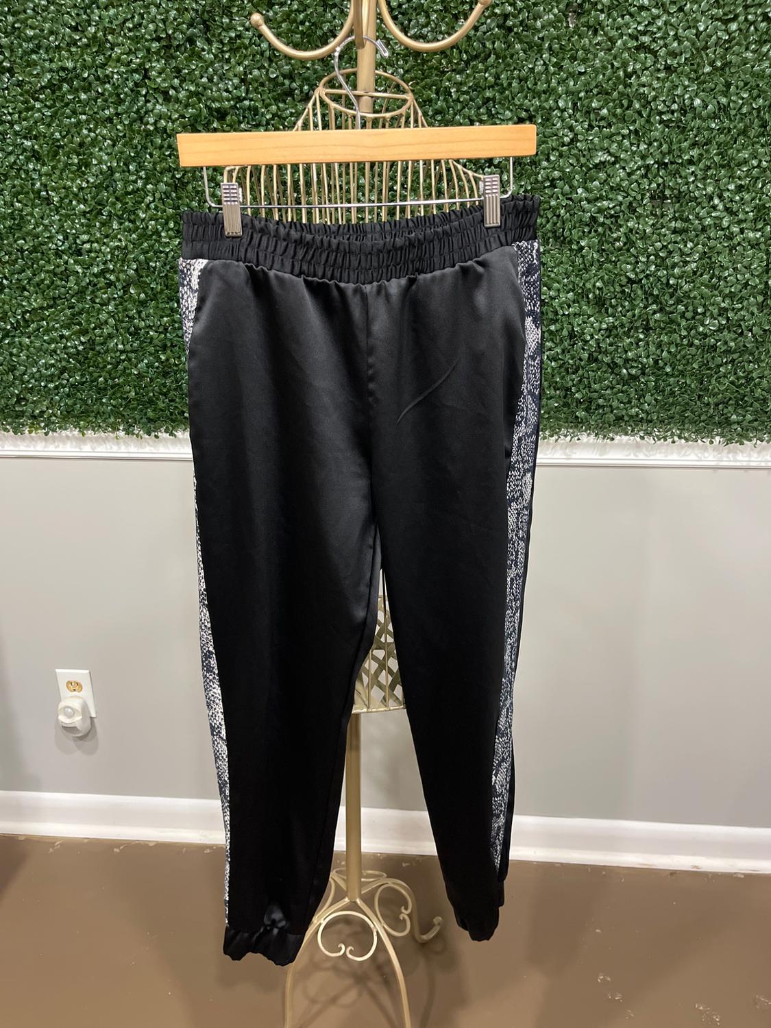 Black pants with an elastic waistband and printed details on the sides, hanging on a clothes rack in front of a green leaf background.