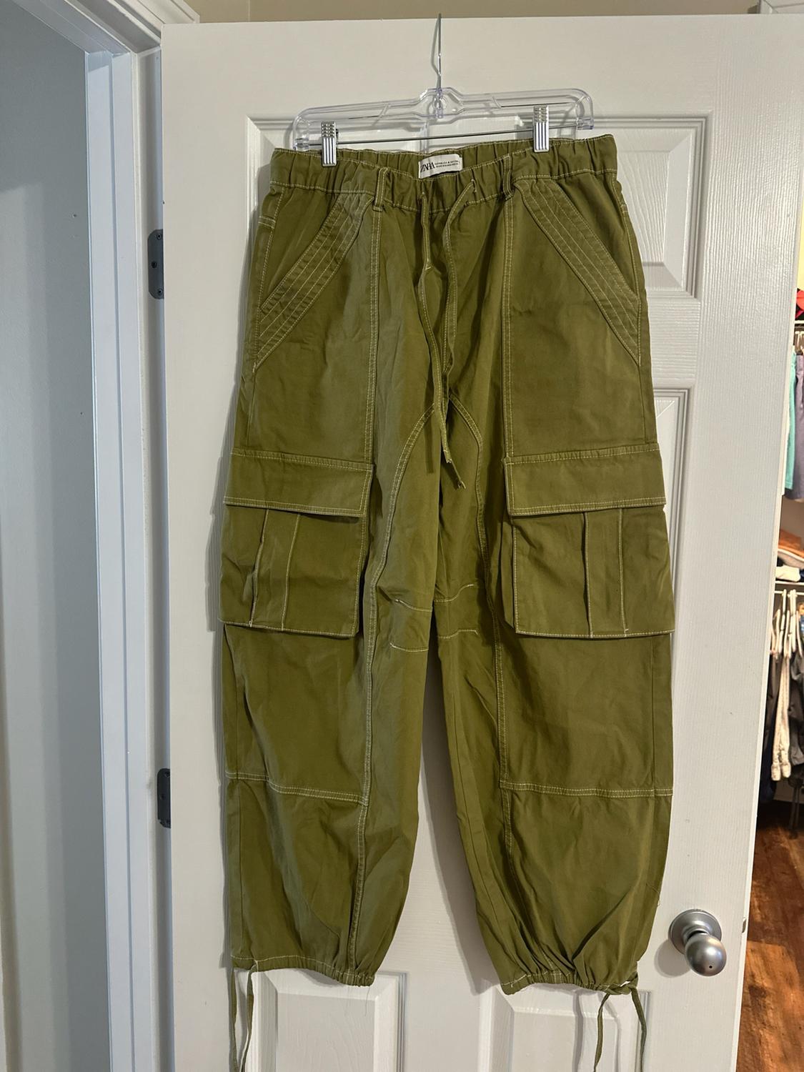 Olive green cargo pants with a ZARA tag, white stitching, an elastic waistband, and pockets, hanging on a white door.