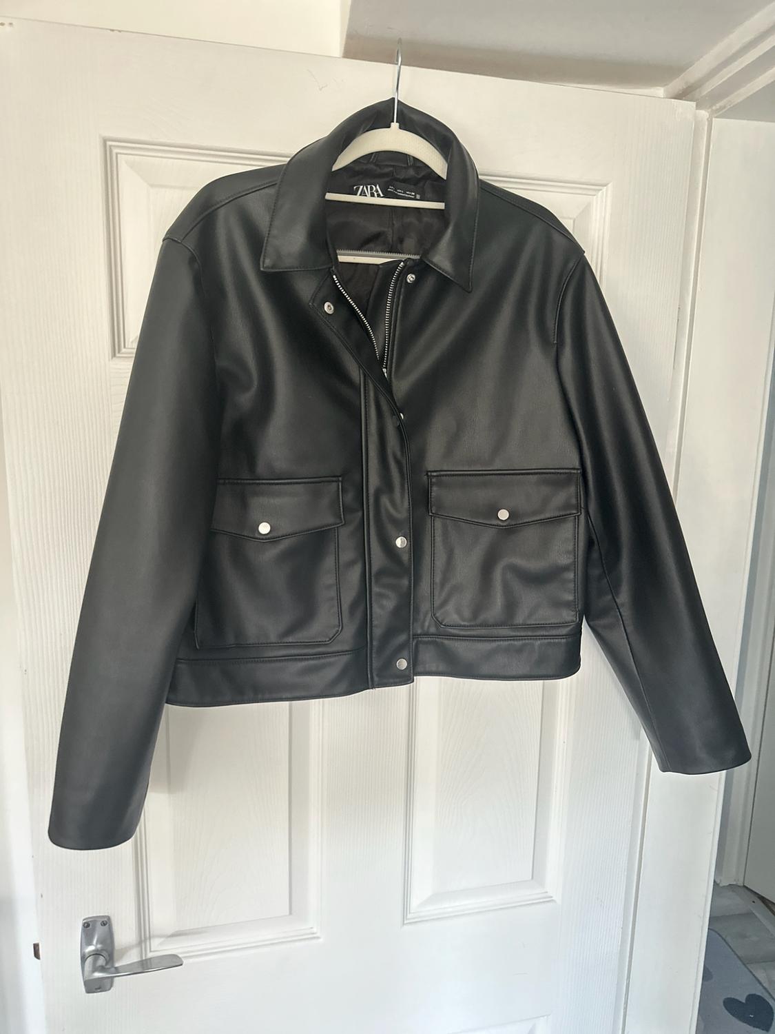 Black jacket hanging on a white door. It has a collar, front fastening, silver buttons and two flap pockets. Label with the text ZARA.