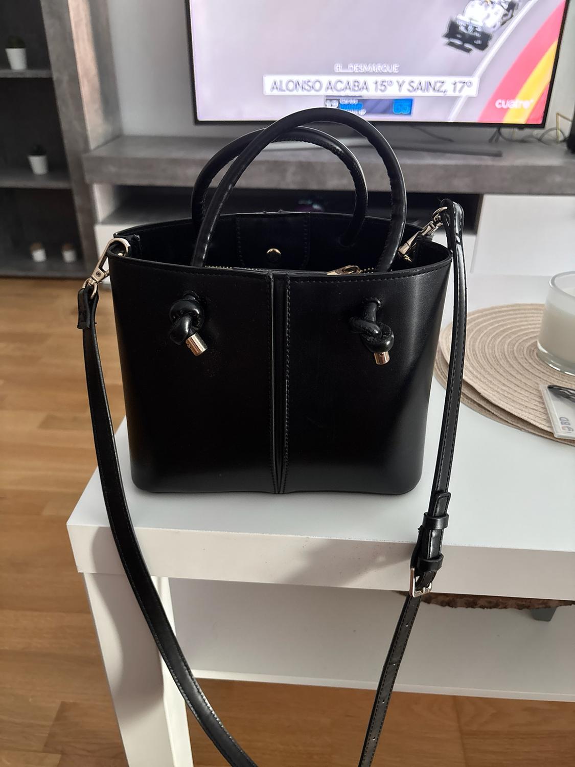 Black handbag with handles and knots on a white table. On the TV: EL DESMARQUE ALONSO FINISHES 15TH AND SAINZ, 17TH cuatro.