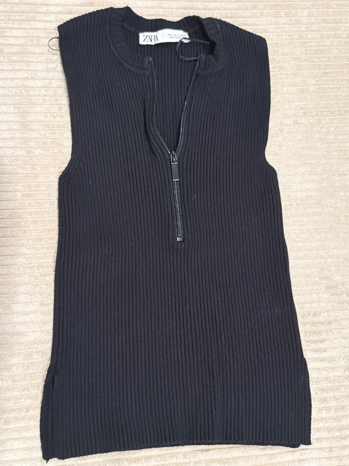 Black sleeveless ribbed knit top with a half-zip front against a textured beige background. Tag with ZARA text.